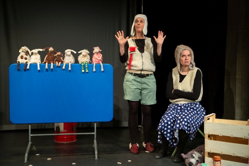 Zwei Personen mit Fellmützen, stehen neben einem blauen Gartentisch auf dessen Kante sieben Schafstofftiere sitzen.