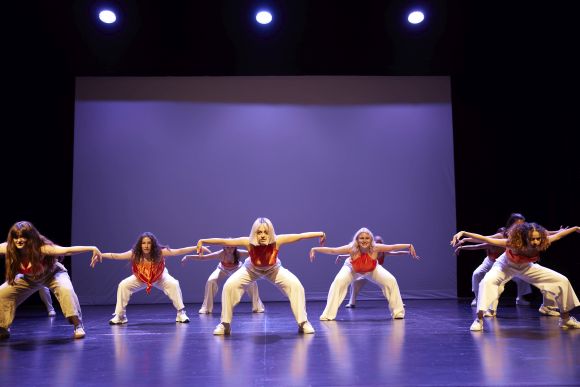 Tänzerinnen auf der Bühne,  in weissen Hosen und orangenen Oberteilen,  mit vorgeneigtem Oberkörper und ausgesterckten Armen.