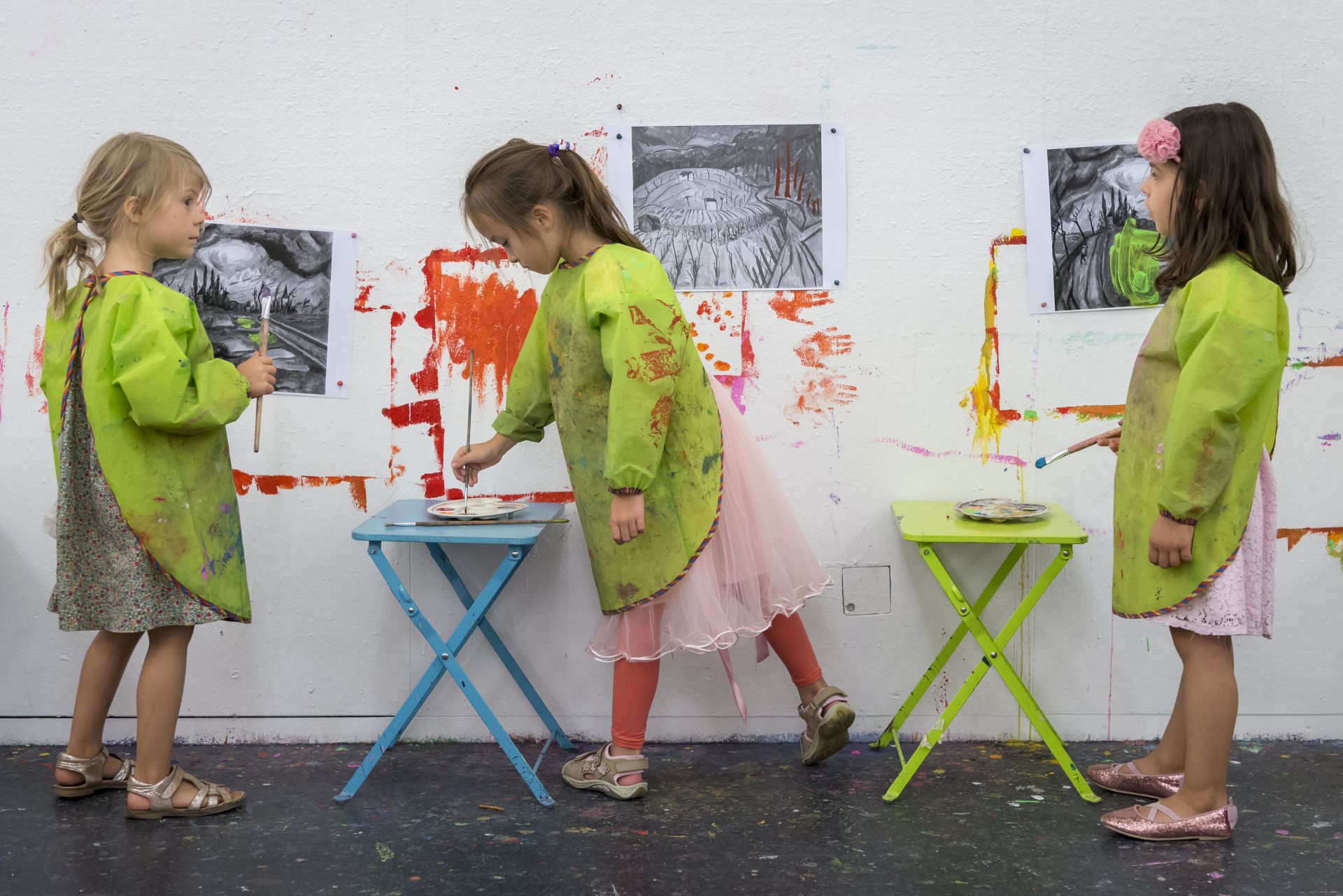Drei Mädchen stehen in grünen Schürzen vor einer Wand und malen