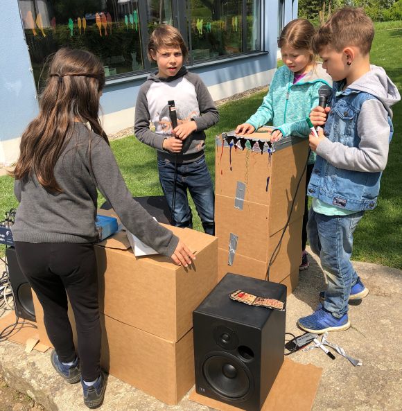 Kinder stehen an Kartonschachteln mit einem Miukrofon und einem Lautsprecher