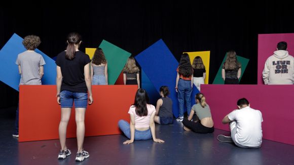 Acht Personen stehen und vier Personen sitzen auf einer Bühne verteilt vor farbigen Stellwänden.