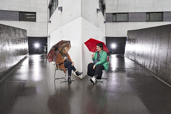 zwei Frauen sitzen mit Regeschirmen vor einem grauen Gebäude
